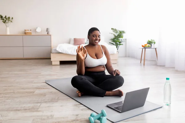 Mujer negra con sobrepeso sentada en una esterilla de yoga frente a una computadora portátil, saludando a un entrenador personal, saludando a la webcam, espacio para copiar —  Fotos de Stock