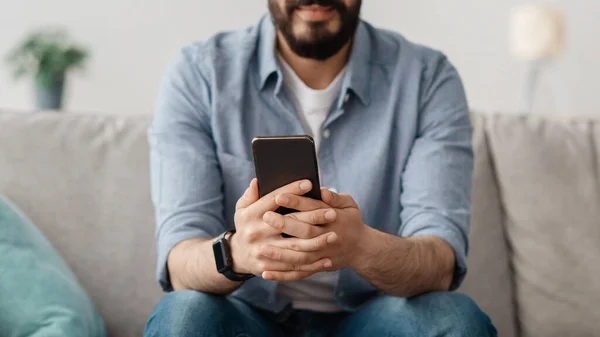 Szoros kép arab férfi használ okostelefon otthon, ül a kanapén, srác böngészés új app vagy üzenetküldés, szelektív fókusz — Stock Fotó