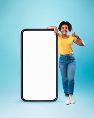 Siyah kadın cep telefonuna boş beyaz ekranla yaslanıyor, baş parmağını kaldırıyor, yeni uygulamalar ya da web sitesi öneriyor,