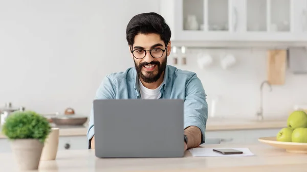 Fjärrkontroll. Glad arabisk frilansare man som arbetar med bärbar dator i köket, skriva på datorn, njuta av online-jobb — Stockfoto