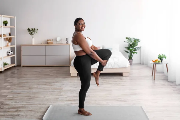 Mujer negra con sobrepeso estirando la pierna, haciendo ejercicios de flexibilidad en la esterilla de yoga, llevando un estilo de vida activo durante el covidio —  Fotos de Stock