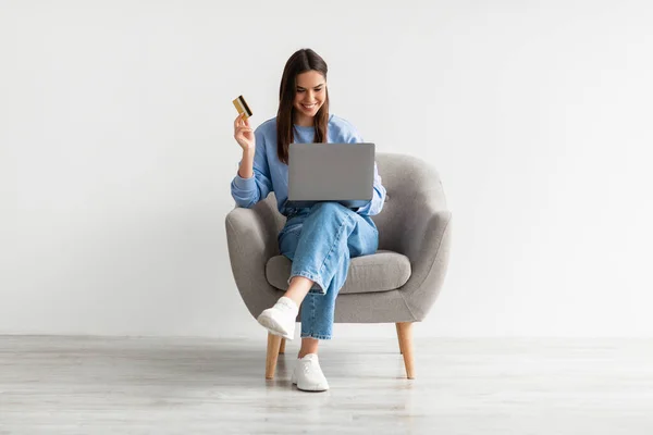 Achats en ligne, e-commerce, services bancaires à distance. Jeune femme assise dans un fauteuil avec ordinateur portable et carte de crédit, acheter sur le web — Photo