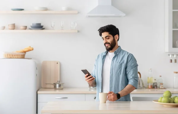 快乐的阿拉伯年轻人用智能手机，在厨房里喝着早晨的咖啡，空荡荡的空间 — 图库照片