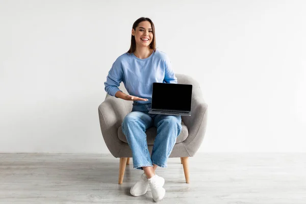 Glad ung dam sitter i fåtölj med bärbar dator, visar tom skärm med mockup, reklam din webbplats design — Stockfoto