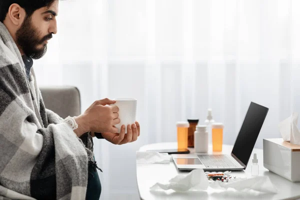 Tratando gripes e resfriados em casa. árabe cara ter febre envolto em cobertor e beber chá quente, assistindo filme no laptop — Fotografia de Stock