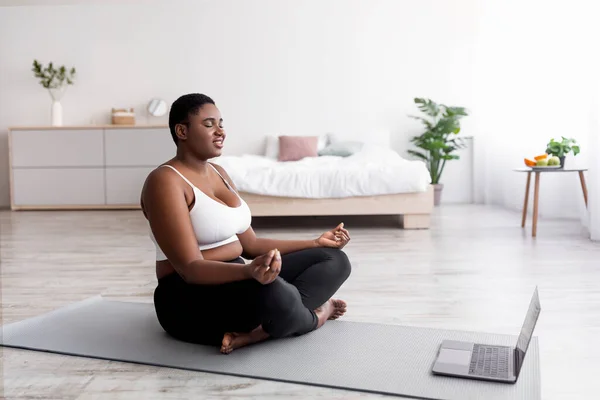 Mujer negra regordeta que tiene meditación en línea o clase de yoga en el ordenador portátil, sentado con los ojos cerrados en la pose de loto en casa —  Fotos de Stock