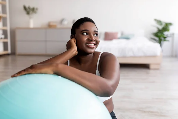 Веселая толстая черная женщина, опирающаяся на фитнес-мяч, занимающаяся спортом в помещении — стоковое фото