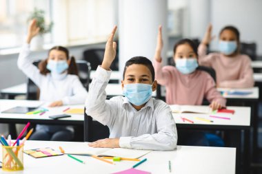Okul çocukları sınıfta el kaldırıyor, koruyucu tıbbi maskeler takıyorlar.