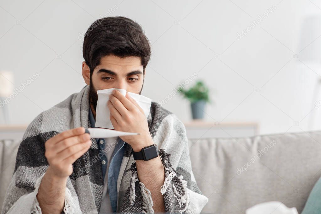 Cold, flu, coronavirus concept. Sick arab man sitting on couch and sneezing, measuring body temperature, home interior