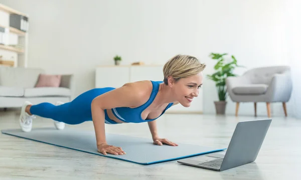 Vrolijke vrouw doen plank op mat met laptop thuis — Stockfoto