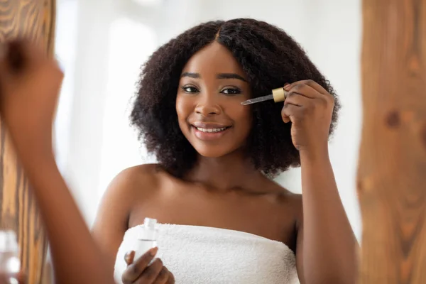 Souriant jeune femme noire en serviette en utilisant du sérum facial près du miroir, prenant soin de sa beauté à la maison — Photo