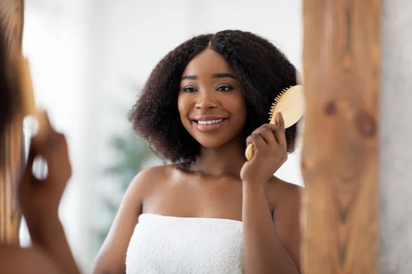 Veselý Afro žena kartáčování její kudrnaté vlasy s dřevěným štětcem před zrcadlem doma — Stock fotografie