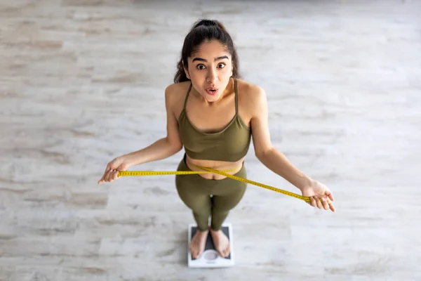 Vor der Ansicht einer schockierten indischen Dame, die auf einer Waage steht und die Taillenmaße überprüft, überrascht mit Ergebnissen der Gewichtsabnahme — Stockfoto