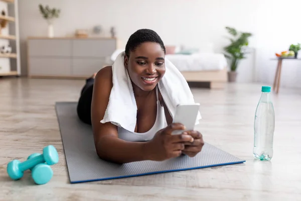 Kurvige schwarze Frau nutzt Smartphone in der Pause vom Training zu Hause, surft im Internet, sucht online nach Workout-Videos — Stockfoto