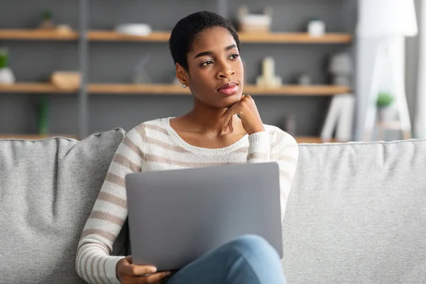 Giovane signora freelancer nero pensieroso seduto sul divano con il computer portatile — Foto Stock