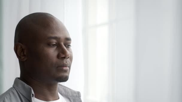 Задумчивый чернокожий мужчина средних лет, стоящий у окна дома — стоковое видео