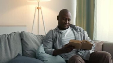 Afro-Amerikan erkek kitap okuyor. Evdeki koltukta oturuyor.