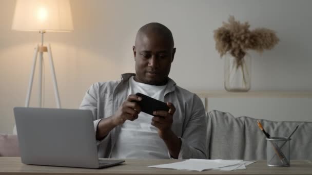 Glada svart man med hjälp av smartphone spela spel online Sitter inomhus — Stockvideo