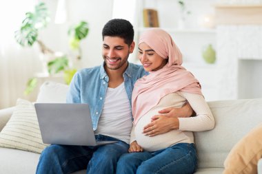 Hamile Müslüman çifti sevmek, bebek odası için mobilya seçmek, oturma odasındaki kanepede dizüstü bilgisayar kullanmak, boş alan.