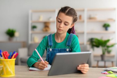 Öğrenci masada oturuyor, dijital tablet kullanıyor, deftere yazıyor.