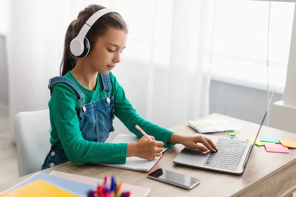 Szorgalmas lány ül az asztalnál, laptopot használ, tankönyvbe ír — Stock Fotó
