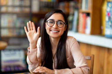 Görüntülü arama. Mutlu genç Arap bayan birini selamlıyor, sanal görüşme yapıyor, kameraya el sallıyor ve gülümsüyor.