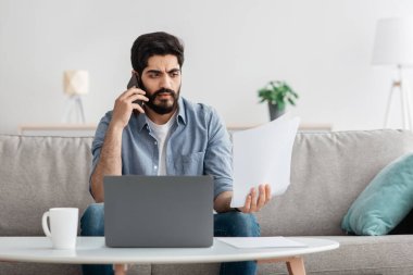 Evden çalışan sinirli Arap işadamı, dizüstü bilgisayarla koltukta oturuyor, belgeleri tutuyor ve telefon görüşmesi yapıyor.