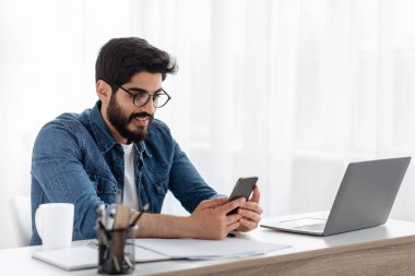 Arap serbest çalışan adam ev ofisinin bilgisayarında çalışırken cep telefonu kullanıyor, yeni uygulamalara veya mesajlara bakıyor.