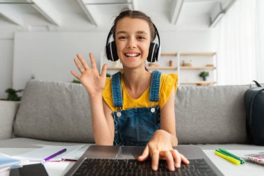 Kulaklıklı kız dizüstü bilgisayar kullanıyor, web kamerasına el sallıyor.