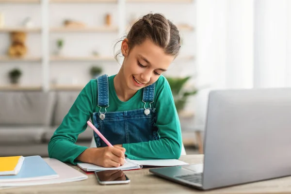 Улыбающийся ученик сидит за столом, пользуется ноутбуком, пишет в блокноте — стоковое фото