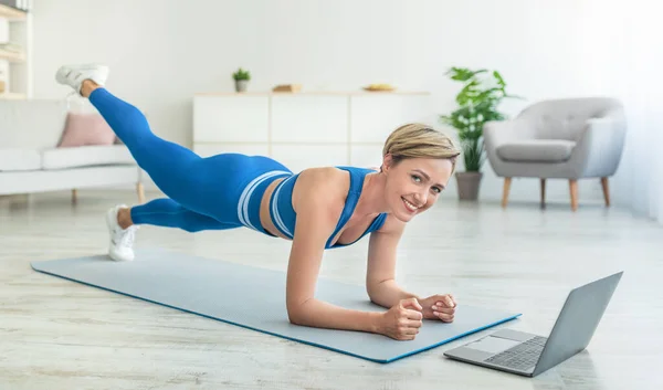 Happy woman doing plank on mat with laptop at home — Stock Photo, Image