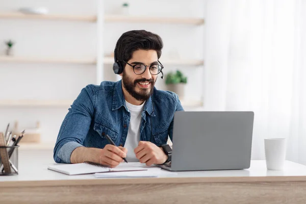 Fernstudium. Glücklicher arabischer Mann mit drahtlosen Kopfhörern, der am Schreibtisch sitzt, Laptop benutzt und in Notizbuch schreibt — Stockfoto