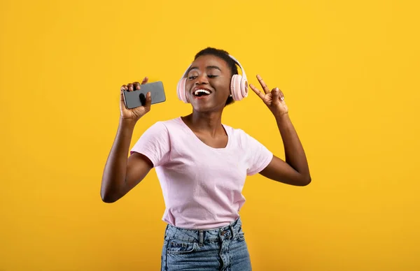 Neşeli Afro-Amerikan bayan akıllı telefona şarkı söylüyor ve sarı arka planda kablosuz kulaklıkla müzik dinliyor. — Stok fotoğraf