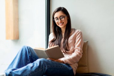 En sevdiğim hobi. Olumlu Arap kadın kafede favori kitabı okuyor, pencerenin yanında oturuyor, boş yer var