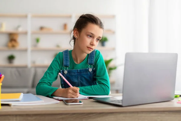 Diligent student zit aan tafel, met behulp van laptop, schrijven in notebook — Stockfoto