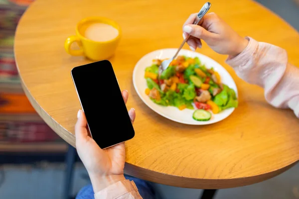 Концепция здорового питания и планирования диеты. Женщина использует смартфон с черным экраном и ест свежий салат в кафе — стоковое фото