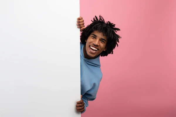 Attractive African American teenager hiding behind blank poster with copy space on pink studio background, mockup — Stock Photo, Image