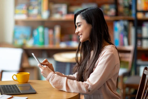 Mujer latina feliz sosteniendo el teléfono móvil y utilizando el ordenador portátil, freelancer trabajando en la cafetería, espacio para copiar, vista lateral —  Fotos de Stock