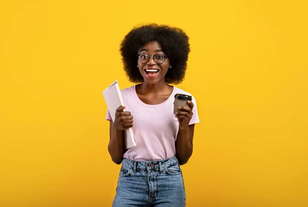 Aufgeregte afrikanisch-amerikanische Studentin hält Notizbücher und Coffee to go in der Hand, Pause, gelber Hintergrund — Stockfoto