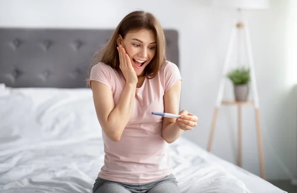 Eccitato giovane donna controllando il suo recente test di gravidanza, sentendosi felice per la futura maternità a letto a casa — Foto Stock