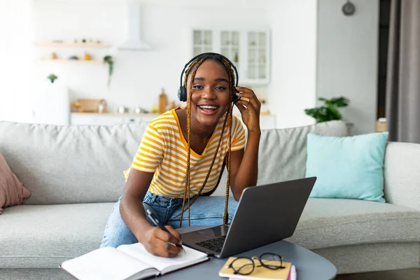 Pozytywny czarny kobieta z słuchawki robi notatki, za pomocą laptopa — Zdjęcie stockowe