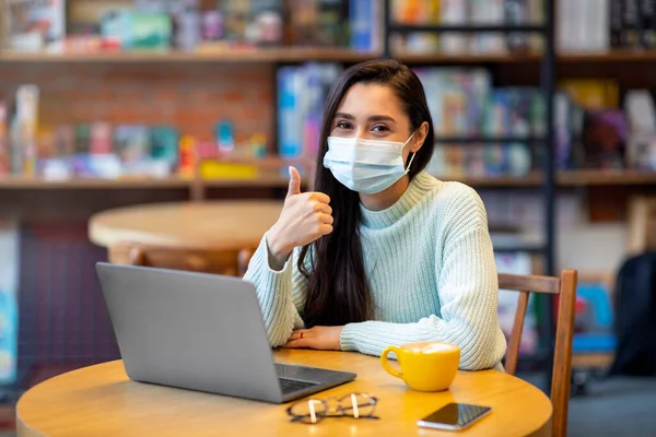 Всё в порядке. Портрет счастливой женщины в хирургической маске, работающей на ноутбуке в кафе, показывающей большой палец вверх — стоковое фото