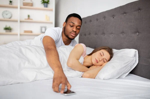 Celos, infidelidad, problemas de relación. Sospechoso marido negro comprobar durmiendo esposas teléfono móvil en la cama — Foto de Stock