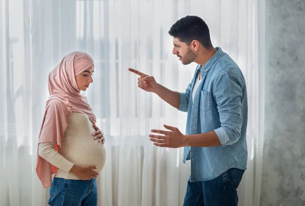 Problemas de relación. Hombre árabe agresivo discutiendo con su esposa musulmana embarazada molesta en casa, de pie cerca de la ventana — Foto de Stock