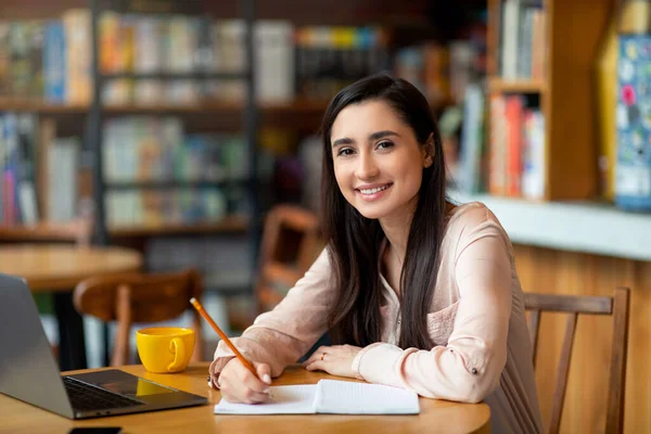 Enthusiastic student latin lady looking for job online, using laptop and taking notes, sitting at cafe, copy space