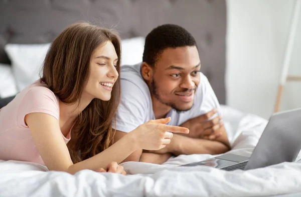 Glada tusenåriga multinationella par ligger på sängen med laptop, titta på roliga video tillsammans — Stockfoto