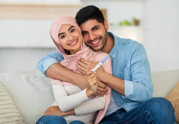 Szerető arab pár bemutató pozitív terhességi teszt ül a kanapén otthon, és mosolygós a kamera — Stock Fotó