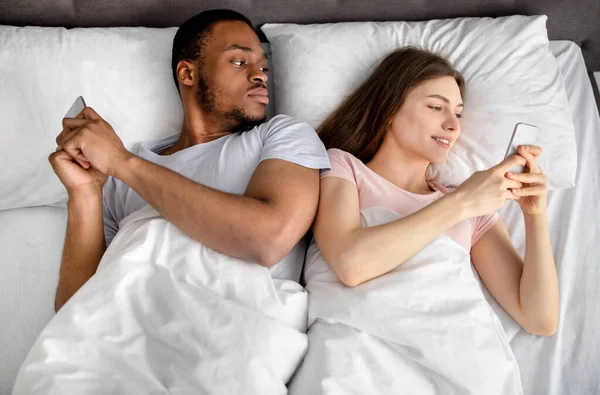 Jeune couple interracial couché dans le lit coincé dans les smartphones, jouer à des jeux en ligne, navigation sur les médias sociaux, vue d'ensemble — Photo