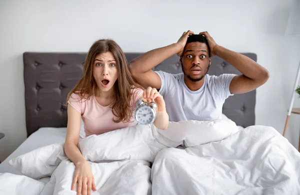 On est en retard. Couple interracial choqué avec réveil saisissant leur tête dans la terreur, assis sur le lit à la maison — Photo