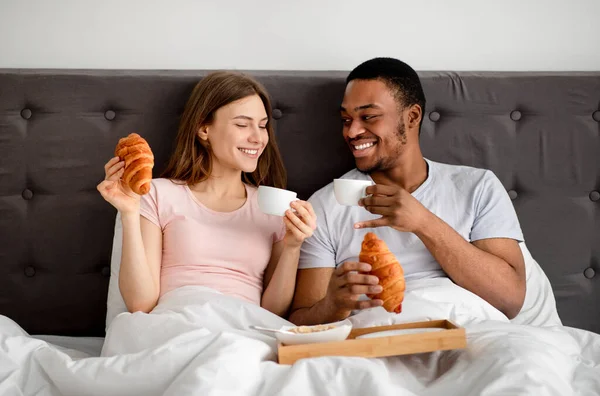 Coppia multirazziale che fa colazione a letto insieme, beve caffè con croissant, trascorre la luna di miele in hotel — Foto Stock